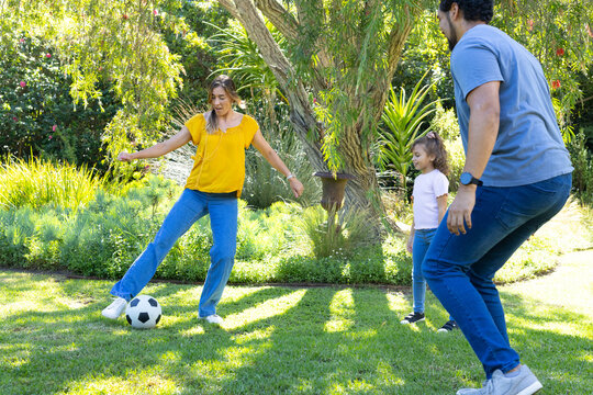 Parents with child playing soccer on backyard lawn, woman in yellow top kicking ball