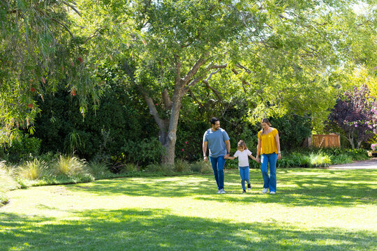 Diverse family of three walking across sunlit grassy lawn under large shade tree, holding hands