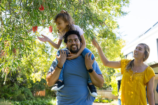 Family man carrying child on shoulders while woman in yellow reaching bottlebrush branch near house