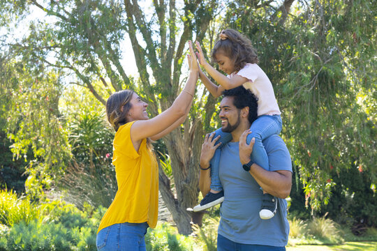 Diverse family highfiving in park, mother in yellow reaching, dad carrying child on shoulders watch