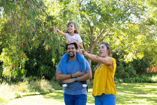 Family trio standing in sunlit park, man carrying child, woman pointing at red flowers
