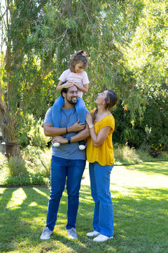 African American family playing under drooping tree on grassy lawn, man carrying child on shoulders
