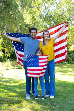 Diverse family standing on sunlit lawn holding large American flag while child waving small flag