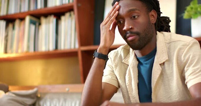 African American man rubbing temples and closing eyes on couch, self-led, bookshelf visible