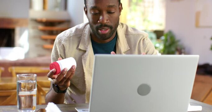 Adult African American man checking pill bottles after reading laptop instructions at dining table