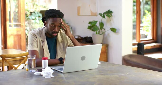 African American man reading urgent email on laptop at home, rubbing forehead, pill bottle, calming