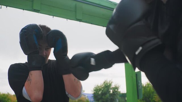 young caucasian boxer practicing defensive sparring under green pavilion with coach mitts, gloves up, concentrated gaze, cloudy sky, slow motion blocks and counter drills in outdoor park