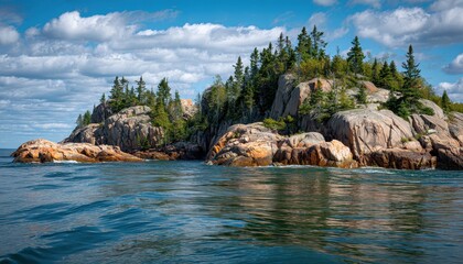 Obraz premium Rugged rocky island with evergreen pine trees surrounded by deep blue ocean water under a bright sunny sky with scattered white clouds in a scenic coastal landscape photography shot