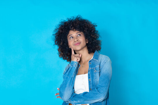 Young afro-latina woman with a thoughtful expression, natural Afro hair, denim jacket, vibrant blue background, concept of doubt, idea and reflection
