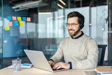 Fototapeta na wymiar Smiling man receiving or providing excellent customer service, wearing a headset and typing on a laptop, working remotely or in a call center office environment