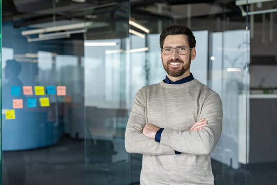 Confident business man wearing glasses and a sweater, standing with arms crossed and smiling broadly in a modern office with glass walls and sticky notes