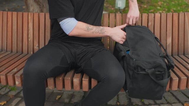white man pulling camera from backpack photographer role helmet on bench tattooed forearms wooden slatted seat cobblestone path daylight urban park careful hands adjust lens and strap ready for street