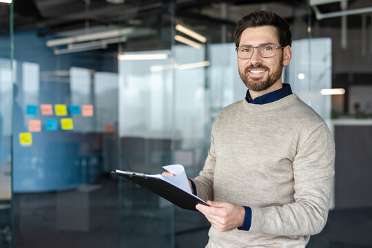 Smiling professional man wearing glasses holding a clipboard and looking at camera, standing in a contemporary office setting with glass walls and colorful sticky notes in the background