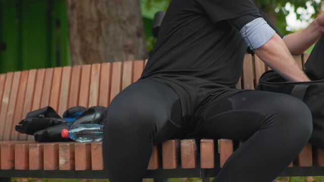 white man opening bag on bench pulls helmet from inner compartment, hands steady fastening chin strap, water bottle and gloves beside him, focused commuter preparing ride in soft daylight