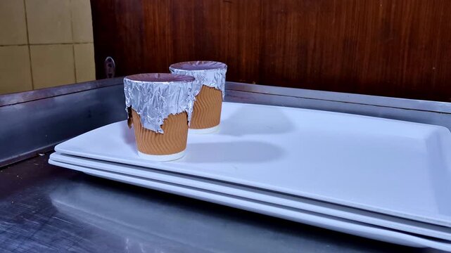 Two small paper cups sealed with crinkled foil lids rest on a stack of rectangular white plates placed on a stainless steel counter against a tiled and wooden backdrop.