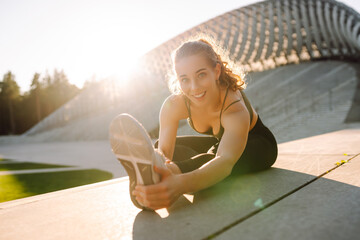 Fototapeta premium An athletic woman exercises on the steps on a sunny day. A young woman in athletic clothing stretches while exercising outdoors at sunset. Concept of activity, sports, and weekends.