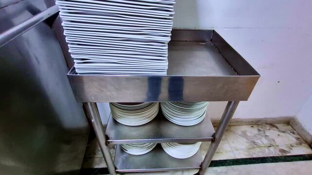A neatly arranged stack of rectangular white plates sits atop a stainless steel rack, with rows of round plates organized on the lower shelves against a plain kitchen wall.