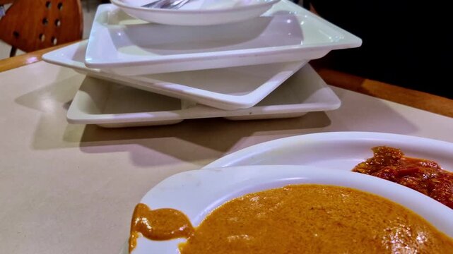 A slightly angled close-up captures a rich, buttery curry in a white bowl with a side of chutney and neatly sliced cucumber, onion, and tomato, while a stack of plates, cutlery, napkins.