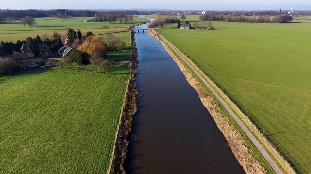 Aerial view of a scenic European river winding through green farmland, alongside a peaceful village featuring a historic brick church with a modern glass nave and houses with solar panels.