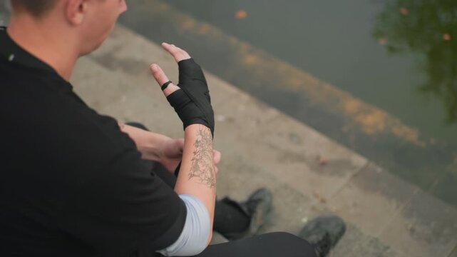 white male taping wrists on steps, tattooed arms, black wrap and slow precise movements, waterfront view meditative athlete preparing, boxing trainee warming, martial artist ritual, tattoo model
