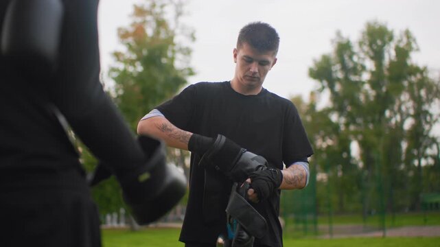 white man facing sparring partner in park, tense stare, hands wrapped inside gloves, measuring distance, focused posture, footwork ready, tattooed arms, dusk greenery, competitive yet respectful