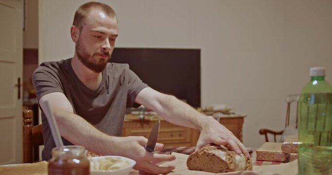 A young man sits at a home table, cutting a piece of fresh bread to accompany his meal. This domestic scene portrays a quiet, everyday moment of preparing and enjoying food.