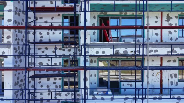 Scaffolding surrounds a building under construction with patches of plaster on the walls. Windows reflect the surroundings on a bright day. Workers can be seen on the site.