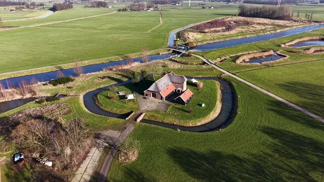 High angle drone view of a straight Dutch canal cutting through vibrant green pastures, featuring a traditional farmhouse, a small water control gate, and a parallel cycling path.