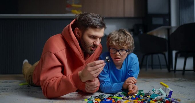 Father and young son lying on the living room floor, concentrating together as they sort colorful building bricks during playful family bonding time