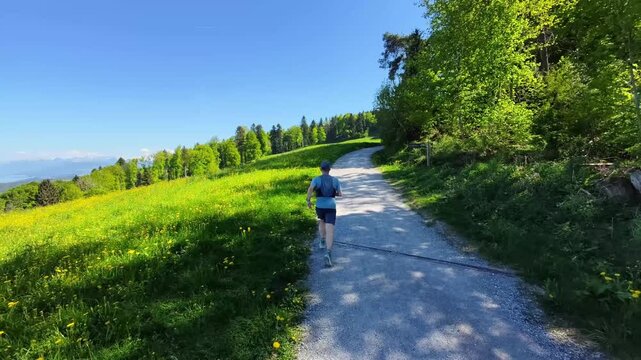 Trail runner from behind crossing open fields in the Swiss countryside, embracing freedom, wide open views and silence, finding mental clarity, balance and a liberating escape from digital noise.
