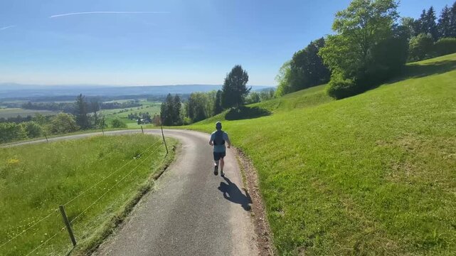 Trail runner from behind crossing open fields in the Swiss countryside, embracing freedom, wide open views and silence, finding mental clarity, balance and a liberating escape from digital noise.