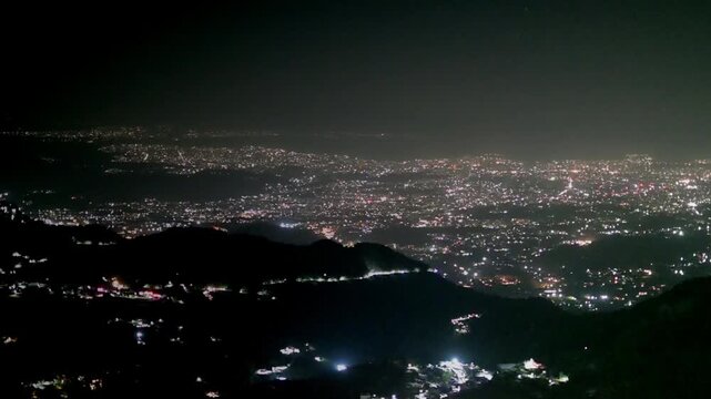 A dazzling night panorama from Mussoorie reveals an expansive valley blanketed with countless twinkling city lights, winding roads, and dark mountain silhouettes under a hazy sky.