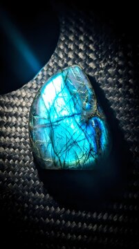 Polished labradorite gemstone displaying brilliant blue flash and iridescent colors on dark woven textile background for luxury jewelry and mineral collection designs.