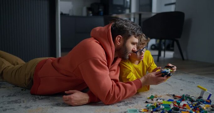 Father and young son lying on living room floor building colorful toy blocks together, sharing focused playtime and creative learning at home