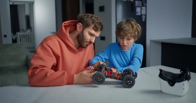 Father and young son repairing a remote controlled toy car together at a kitchen table, exploring mechanics and technology with VR headset nearby
