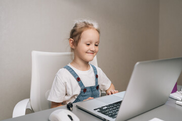 Cute smiling child studies online using laptop at home. Girl sitting at desk engaged in distance...