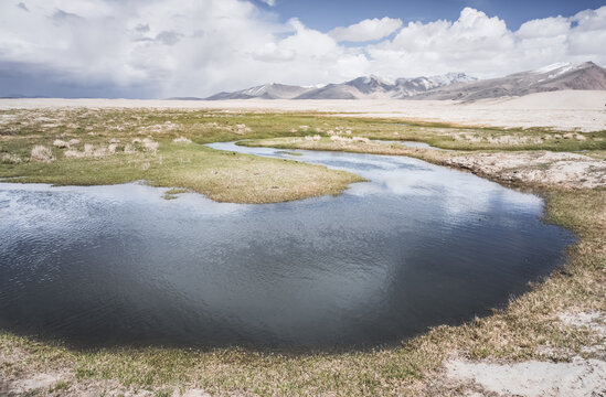 High in the Pamir Mountains of Tajikistan, a river flows along a raft with grassy banks against a backdrop of snow-capped mountain peaks. A panoramic view of the Tien Shan highlands