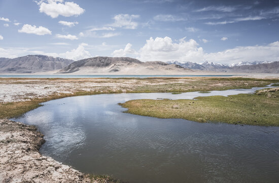 High in the Pamir Mountains of Tajikistan, a river flows along a raft with grassy banks against a backdrop of snow-capped mountain peaks. A panoramic view of the Tien Shan highlands