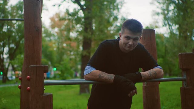 white male resting on pullup bar in urban park at dusk, tattoos and workout gloves visible, leaning on metal rail between sets, thoughtful expression, soft green trees background, gritty yet calm