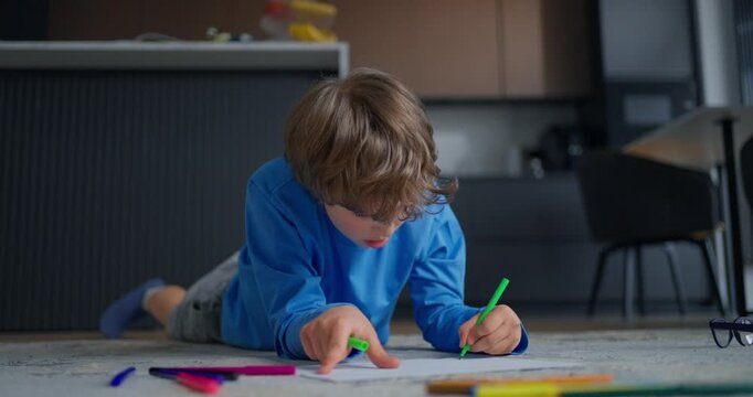 Young boy lying on carpeted floor drawing with colorful markers in a modern home interior, focused on creative play and childhood learning