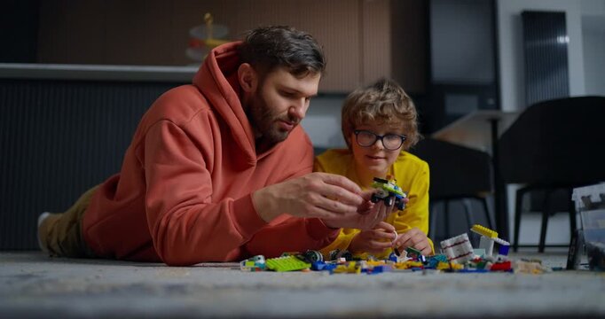 Father and young child lying on living room floor building colorful toy blocks together, sharing focused playtime and creative learning at home