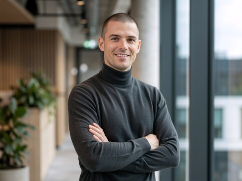 Confident man with arms crossed in modern office hallway. Smiling professional in grey turtleneck sweater. Modern business portrait in contemporary workplace