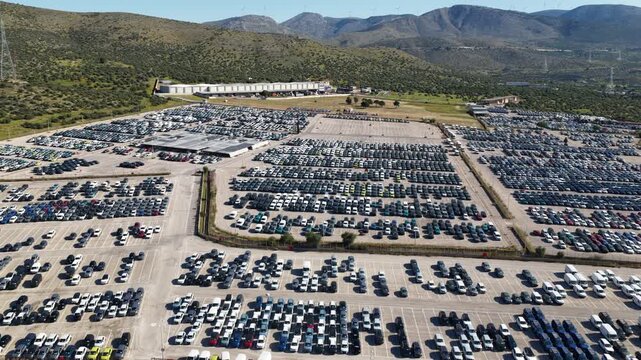 Vast aerial perspective showing thousands of new cars neatly arranged in a massive distribution hub parking lot, awaiting export or delivery to dealerships within a large industrial complex