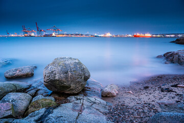 Large coastal boulder facing brightly lit harbor cranes and cargo ships long exposure. © Trygve