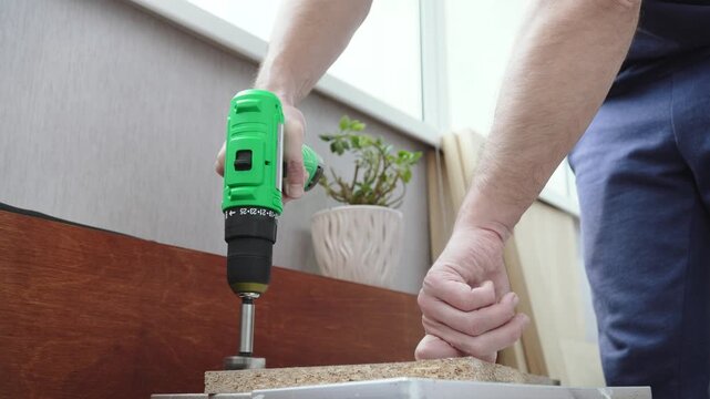Caucasian man drilling hole with forstner bit using screwdriver to install hinges closeup. Drilling furniture parts made of chipboard during manufacturing in workshop.