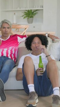 Vertical video: Running man in white tee sitting on floor at home with green beer cheering at match
