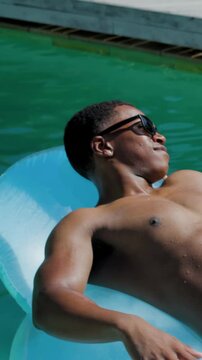 Vertical video: Sunlight drawing man on blue float turning toward dock in pink trunks dark shades