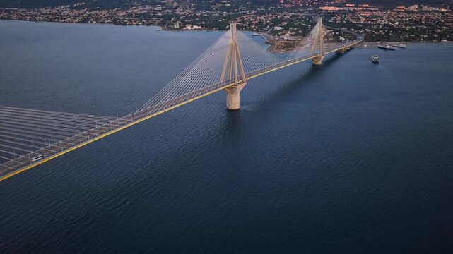Magnificent aerial view showing the illuminated Rio-Antirrio bridge crossing the gulf of Corinth in Greece, with traffic moving across the span and the coastal city visible at dusk