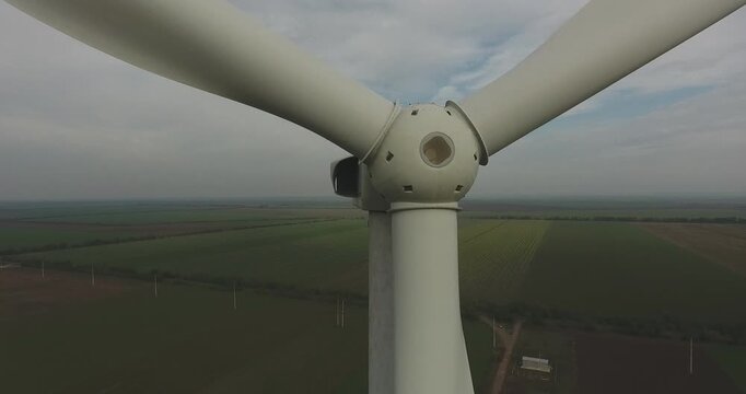 Large wind turbine blades rotate above expansive agricultural fields under cloudy sky conditions. Industrial renewable energy infrastructure dominates the rural landscape with visible mechanical motio