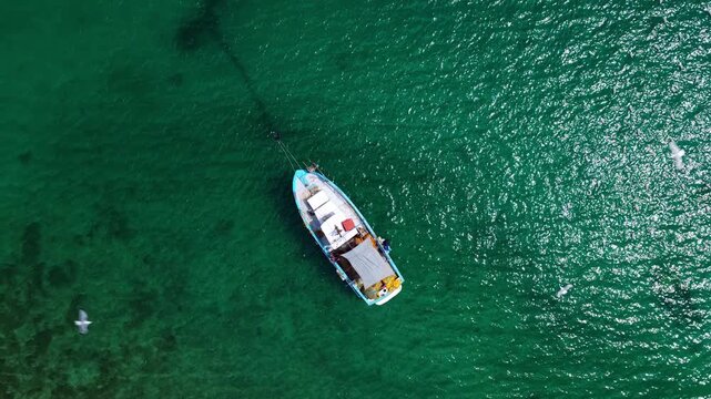 An overhead drone ascends, widening the frame from a solitary traditional fishing boat anchored in vibrant emerald water. As it rises, a flock of seagulls is revealed circling and around the vessel